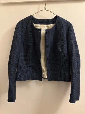 Mörkblå prickig kavaj - Snygg mörkblå kavaj med små vita prickar. Rak passform, rundad halsringning och helknäppning med knappar framtill. Fodrad insida och två fickor fram. Perfekt för en clean och stilren look.