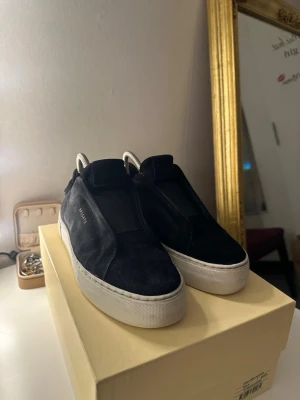 Svarta sneakers från Axel Arigato - Stilrena svarta sneakers från Axel Arigato i mocka med vit platt sula. Skorna har minimalistisk design utan synlig snörning och diskret guldfärgad logga på sidan. Perfekta för dig som gillar en clean och modern look.