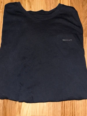 Svart t-shirt från GANT i bomull - En stilren svart t-shirt från GANT i mjuk bomull. Klassisk passform med rund halsringning och diskret GANT-logga på bröstet. Perfekt för en clean och enkel look. Använd en gång.