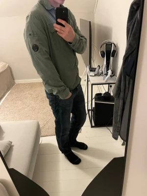 Grön overshirt från C.P. Company - Säljer en stilren grön overshirt från C.P. Company. Jackan har klassisk krage, bröstficka med lock och ikonisk linsdetalj på ärmen. Tillverkad i bomull och har dragkedja framtill. Perfekt för lager-på-lager och snygg till jeans.