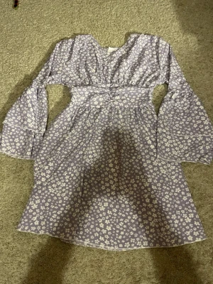 Ljuslila blommig klänning - Supersöt ljuslila klänning med vita blommor och v-ringning. Klänningen har långa, vida ärmar och markerad midja som ger en fin siluett. Perfekt för vår och sommar! Materialet känns lätt och luftigt.