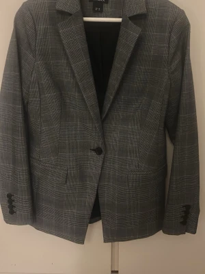 Rutig kavaj från Lindex, grå - Snygg rutig kavaj från Lindex i grå toner, storlek 38. Klassisk modell med en knapp framtill, fickor och långa ärmar. Tidlös design som passar till många olika looks. Fodrad insida och extra knappar medföljer.