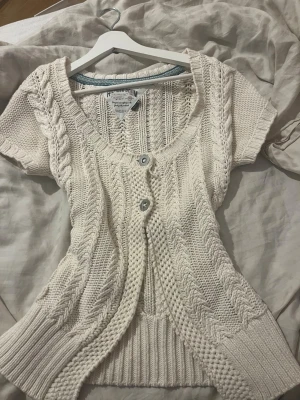 Vintage Vit kortärmad kofta Hilfiger Denim - Kortärmad kofta från Hilfiger Denim i tjockt, stickat material. Färgen är krämvit och koftan har flätstickade detaljer, två knappar framtill och ribbade muddar. Perfekt för lager på lager och chill vår- eller höststil.