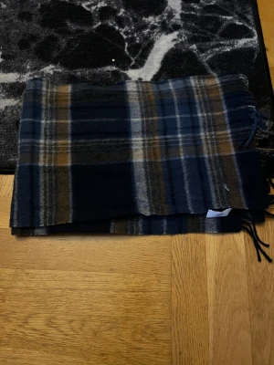 Rutig ullväst från Bläck - Säljer en rutig väst från Bläck i mjuk ullmix. Västen har mörkblå, bruna och grå toner med klassiskt rutmönster och fransar i nederkant. Perfekt för dig som vill ha en stilren och tidlös look.