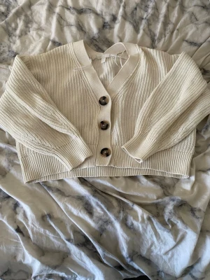 Beige croppad kofta med knappar - Snygg beige croppad kofta med grovstickad struktur och stora bruna knappar framtill. Koftan har v-ringad hals och långa ärmar med ribbade muddar. Perfekt för lager-på-lager och chill stil.