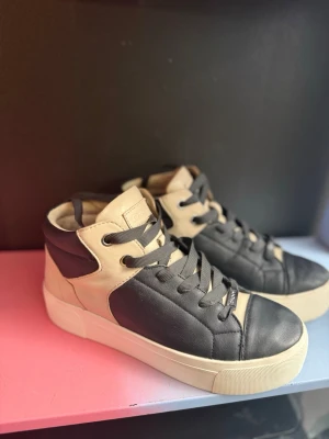 Svart-beige sneakers från DKNY break - Snygga sneakers från DKNY i svart och beige skinn med högt skaft och svarta snören. Klassisk design med rund tå och vit platt sula. Perfekta för dig som vill ha en clean och stilren look till vardags.