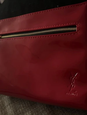 Röd YSL nessesär  - Säljer en glansig röd nessesär från YSL Beauté med guldfärgad dragkedja och ett snyggt läppformat hänge.