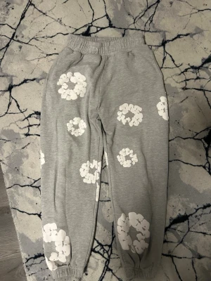 Grå mjukisbyxor med vita blommor - Säljer ett par grå mjukisbyxor med elastisk midja och benslut. Byxorna har stora vita blommiga tryck över hela benen som ger en chill och unik vibe. Perfekta för att hänga hemma eller till vardagslooken.