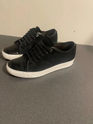 Svarta sneakers med vit sula - Säljer ett par stilrena svarta sneakers med vit sula. Skorna har snörning och en kombination av mocka och glansigt skinn på tån. Klassisk låg modell som passar till det mesta och ger en clean look.