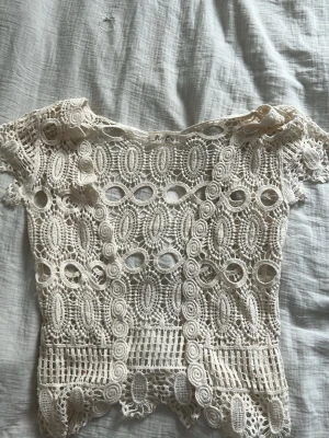 Vit spetsblus från Miss Line - Säljer en superfin vit blus från Miss Line med detaljerad spets och genombrutet mönster. Blusen har korta ärmar och ett unikt virkat mönster med ovala former och vågig nederkant. Perfekt för dig som gillar boho-vibbar och vill sticka ut.