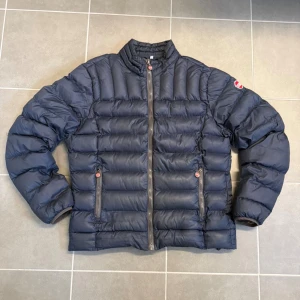 Mörkblå dunjacka från Colmar - Säljer en mörkblå dunjacka från Colmar med klassisk quiltad design och hög krage. Jackan har dragkedja framtill, två sidofickor med dragkedja och Colmar-logga på ärmen. Perfekt för kyliga dagar och riktigt snygg till vardagslooken. Storlek M