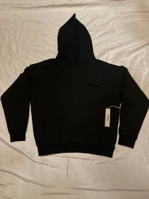 Essentials Hoodie - Svart Essentials hoodie med minimalistisk logga. Klassisk passform med huva och känguruficka. Storlek medium