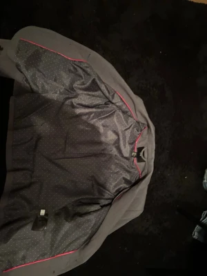 Grå kavaj från H&M med knappar - Snygg grå kavaj från H&M med två knappar framtill och klassisk krage. Fodret är mörkgrått med små prickar och en rosa kant. Kavajen har fickor med lock och fyra dekorativa knappar vid ärmsluten. Perfekt för en clean och stilren look.