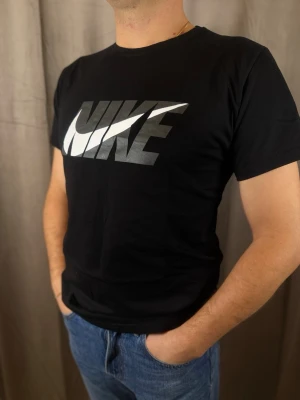 Svart Nike t-shirt Dri-Fit - Svart t-shirt från Nike med stor logga i vitt och grått över bröstet. Klassisk rund hals och korta ärmar. Tillverkad i Dri-Fit material som andas, perfekt för en aktiv livsstil. Enkel och stilren design som funkar till det mesta.