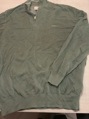 Grön stickad half zip-tröja från GANT - Snygg grön stickad tröja från GANT med half zip och hög krage. Tröjan har diskret GANT-logga broderad på bröstet och ribbade muddar vid ärmslut och nederkant. Perfekt för lager-på-lager och chill stil.