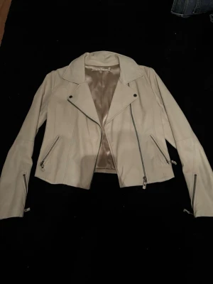 Beige skinnjacka med dragkedjor - Snygg beige skinnjacka med klassisk bikerstil och flera dragkedjor framtill. Jackan har långa ärmar och detaljerade sömmar. Perfekt för dig som vill ha en stilren och edgy look till vardagsoutfiten.