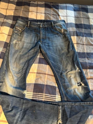 Diesel Krooley blå jeans med slitningar - Säljer ett par blå Diesel Krooley jeans med snygga slitningar och distressed detaljer på både fram- och baksida. Jeansen har klassisk femficksdesign, raka ben och cool faded effekt. Perfekta för dig som gillar en avslappnad och trendig look.