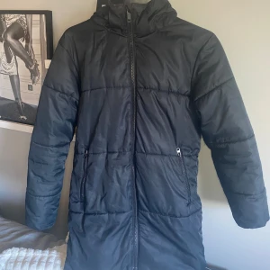 Svart lång pufferjacka Everest 158 - Svart lång pufferjacka från Everest med huva och två sidofickor med dragkedja. Jackan har quiltad design och hel dragkedja framtill. Perfekt för kalla dagar och har en clean look som funkar till allt. Snygg och enkel att matcha med din stil.