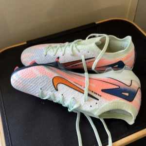 Nike Air Zoom Mercurial fotbollsskor - Säljer ett par Nike Air Zoom Mercurial fotbollsskor i vitt med inslag av ljusrosa och marinblå detaljer. Skorna har orange Swoosh-logga, snörning och en lätt, stickad ovandel. Sulan har tydliga dobbar för bra grepp på planen. Perfekta för dig som vill sticka ut på matchen!