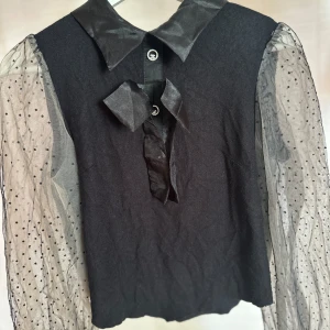 Svart blus med transparenta prickiga ärmar - Snygg svart blus med transparenta, långärmade ärmar i mesh med svarta prickar. Blusen har en glansig krage, rosett framtill och dragkedja i ryggen. Perfekt för dig som vill sticka ut med coola detaljer och trendig look.