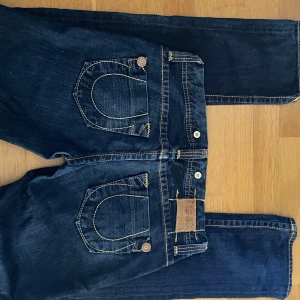True Religion jeans mörkblå, stl 28 - Mörkblå jeans från True Religion med ikoniska kontrastsömmar och stora bakfickor med knappdetaljer. Klassisk femficksmodell med knappgylf och tydlig logga på etiketten. Snygg passform och coola detaljer, perfekta för en streetinspirerad look.