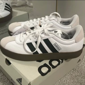 Adidas sneakers  - Snygga Adidas sneakers i vitt med klassiska svarta ränder på sidorna och brun sula. Skorna har snörning, rund tå och är tillverkade i en mix av syntet och mocka. Perfekta för hösten. Köpta för 999:-. Knappt använda