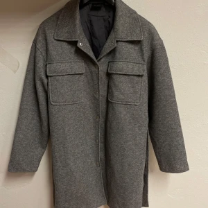 Grå kappa med bälte Vero Moda - Snygg grå kappa från Vero Moda i storlek M. Jackan har stora bröstfickor, knappar framtill och ett bälte i midjan för extra stilpoäng. Långärmad och med klassisk krage, perfekt för lager-på-lager under hösten och våren.