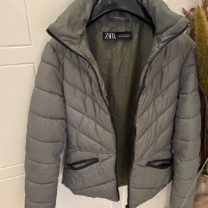 Grå/grön pufferjacka från Zara - Snygg grå pufferjacka från Zara med quiltad design och dragkedja framtill. Jackan har hög krage och två praktiska fickor med dragkedja. Perfekt för kyliga dagar och enkel att matcha med olika outfits.