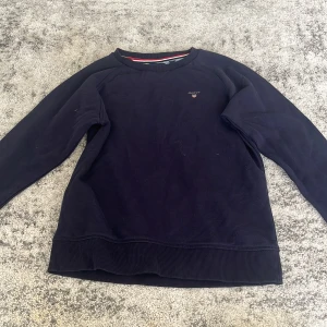 Mörkblå sweatshirt från GANT - Mörkblå sweatshirt från GANT med diskret logga på bröstet. Tröjan har rund hals med snygg randig insida i rött, vitt och blått. Långa ärmar och ribbade muddar vid ärmslut och nederkant. Perfekt för dig som gillar klassisk och enkel stil.