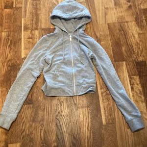 Grå croppad hoodie från H&M Divided - Grå croppad hoodie från H&M Divided med dragkedja framtill och huva. Tröjan har långa ärmar, ribbade muddar och känguruficka. Perfekt för en chill och avslappnad stil. Tillverkad i mjuk bomullsmix.