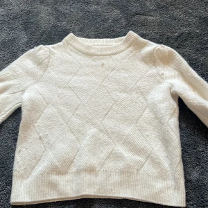 Vit stickad tröja från Dobber - Vit stickad tröja från Dobber med diskret rutmönster och ribbade muddar. Tröjan har lång ärm och en lite högre halsringning, perfekt för kyligare dagar. Enkel och stilren design som passar till det mesta.