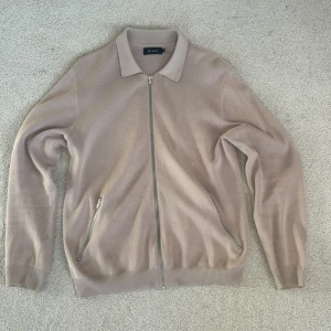 Beige kofta med dragkedja från Riley - Säljer en stilren beige kofta från Riley med hel dragkedja framtill och två fickor med dragkedja. Tröjan har klassisk krage, ribbade muddar och är stickad i mjukt material. Perfekt för lager-på-lager under höst och vår.