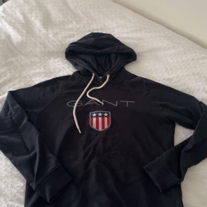 Svart hoodie från Gant med logga XS - Svart hoodie från Gant med broderad logga och sköld på bröstet. Tröjan har huva med vita snören och långa ärmar. Perfekt för dig som gillar klassisk och sportig stil.