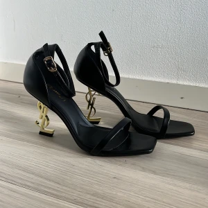 Svarta YSL klackar med gulddetalj - Snygga svarta klackskor med öppen tå och smal rem över foten. Klacken är i guld och formad som ett stiliserat monogram.Skorna har justerbart ankelband med spänne och fyrkantig tå. Tillverkade i syntetmaterial.