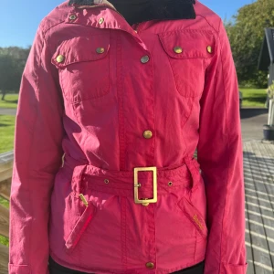 Rosa jacka med bälte från Barbour - Snygg rosa jacka från Barbour med guldiga knappar, bälte i midjan och fyra fickor framtill. Jackan har lång ärm, klassisk krage och rutigt innerfoder i rosa och vitt. Perfekt för dig som vill sticka ut med färg och stil. Ny pris 3500kr
