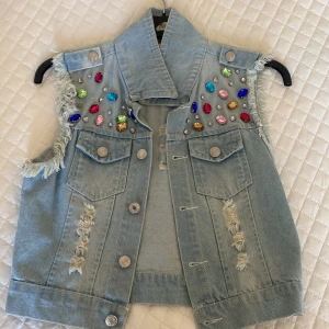 Ljusblå jeansväst med färgglada stenar - Ljusblå jeansväst med slitna detaljer, fransade ärmhål och stora färgglada stenar på axlarna. Västen har två bröstfickor med knappar och en cool, edgy look. Perfekt för dig som vill sticka ut med din stil.