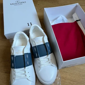 Valentino Garavani Open sneakers vit/blå - Snygga helt oanvända Valentino Garavani Open sneakers i vitt skinn med bred blå läderdetalj över vristen. Kommer med originell låda.  Klassisk rund tå, vita snören och exklusiv känsla. Guldlogga på plösen och cool mönstrad sula. Perfekta för dig som vill sticka ut med stil.
