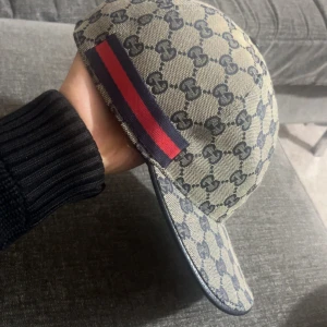 Gucci keps med GG-mönster och rödblå rand - Snygg keps från Gucci i beige och marinblått med klassiskt GG-monogram över hela kepsen. På sidan finns en ikonisk röd och marinblå rand. Kepsen är tillverkad i bomullscanvas och har justerbar rem baktill för perfekt passform.