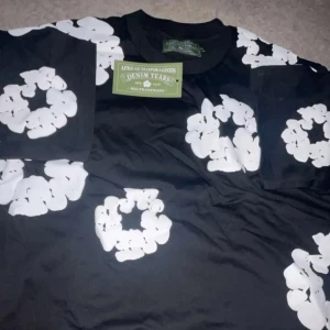 Svart t-shirt Denim Tears med blommor - Svart t-shirt från Denim Tears med stora vita blommiga tryck över hela tröjan. Klassisk rund halsringning och korta ärmar. Tillverkad i mjuk bomull, perfekt för en cool och avslappnad stil.