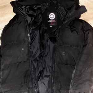 Svart dunjacka från Canada Goose XS - Säljer en svart dunjacka från Canada Goose i storlek XS. Jackan har huva, klassisk Canada Goose-logga på ärmen och dragkedja framtill. Perfekt för kalla dagar med sin tjocka, vadderade design och stilrena look.