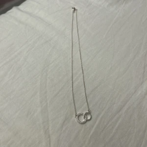 Silverhalsband med två ringar - Stilrent halsband i silver med två sammanlänkade ringar, varav en är dekorerad med små gnistrande stenar och den andra är slät. Perfekt för dig som gillar minimalistiska smycken med en touch av elegans.