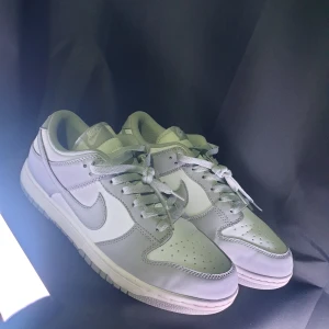 Nike Dunk Low vit/grå sneakers - Nike Dunk Low sneakers i vitt och ljusgrått skinn. Klassisk siluett med rund tå, snörning och platt sula. Swoosh-logga på sidorna och Nike-broderi på hälen. Perfekta för dig som gillar stilrena och cleana sneakers med retrovibe.
