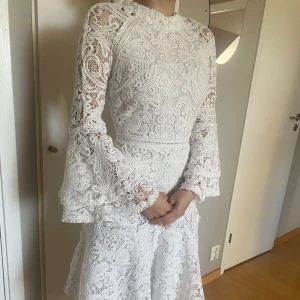 Vit spetsklänning med volangärmar - Superfin vit klänning i spets med långärmade trumpetärmar och vackra volangdetaljer. Klänningen har hög hals, figurnära passform upptill och faller snyggt i kjolen. Perfekt för dig som vill sticka ut med en romantisk och elegant look.