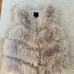 Fluffig pälsväst från New Look - Supermjuk och fluffig pälsväst från New Look i ljusbeige ton. Västen har långa, fejkade pälsstrån som ger en lyxig och trendig look. Perfekt att slänga över en hoodie eller stickad tröja för extra stilpoäng.