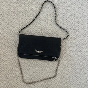 Svart axelväska med kedja och ormmönster - Snygg svart axelväska i skinnimitation med ormmönstrad yta och silverfärgad kedjerem. Väskan har dekorativt metallspänne med vingar framtill och dragkedjor både fram och bak. Perfekt för dig som vill ha en edgy och trendig look. Inget kvitto därför jag säljer biligt