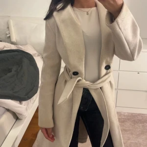 Beige kappa med bälte och knappar - Snygg beige kappa med markerad midja tack vare knytbälte och två stora svarta knappar framtill. Lång modell med klassisk krage och raka ärmar. Perfekt för dig som vill ha en stilren och tidlös look. Priset går att diskutera vid snabbt köp. 