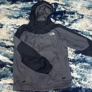 Svart och grå vindjacka från The North Face - Snygg svart och grå vindjacka från The North Face med huva och broderad logga på bröstet och ryggen. Jackan har meshfoder på insidan och är tillverkad i polyester, perfekt för blåsiga dagar. Klassisk passform och stilren design.