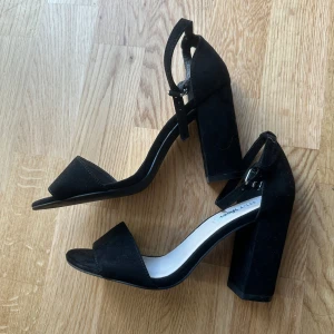 Svarta klackskor från NLY Shoes - Stilrena svarta klackskor från NLY Shoes med bred klack och öppen tå. Skorna har en enkel rem över tårna och justerbar rem runt vristen. Ytan är i mockaliknande material som ger en snygg och modern look. Perfekta för att lyfta vilken outfit som helst.
