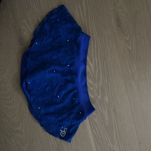 Blå kånståknings kjol  - Supersöt blå kortkjol med blommig spets, små pärlor och glittrande strasstenar i form av ett S. Kjolen har en mjuk satinliknande midja och är perfekt för dig som vill sticka ut med detaljer. Snygg och feminin look!