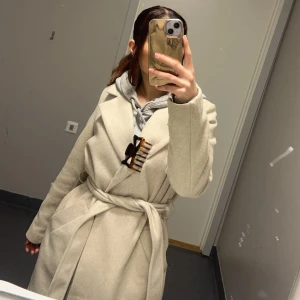 Beige kappa med bälte - Snygg beige kappa med bälte i midjan och breda slag. Jackan har lång ärm och en klassisk, rak passform. Perfekt för dig som gillar stilrena och tidlösa plagg. Materialet känns mjukt och värmande, perfekt för kyligare dagar. Den är lite nopprig men det går att ta bort. 